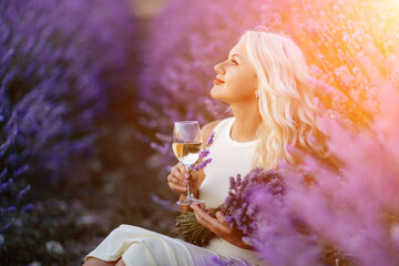 Blonde lavender field holds a glass of white wine in her hands. Happy woman in white dress enjoys...