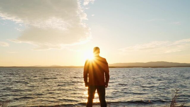 A man stands on the shore of a lake and looks at the sunset, inspired by adventure, success, nature, beauty, achievements and freedom.