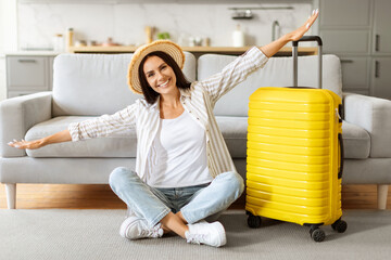 Excited woman in straw hat, sitting with arms wide open beside suitcase