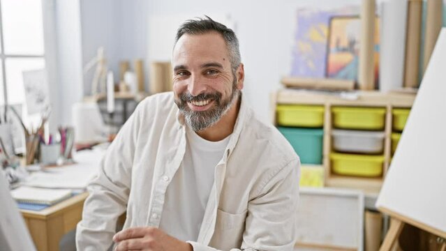 A mature bearded man with grey hair smiles while contemplating a canvas in an art studio.