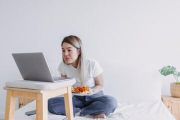 Asian woman eating pasta while watching series from notebook, sitting on white bed and working at home, smiling and looking at camera.
