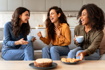 Three joyful multicultural women friends enjoying coffee and cake indoor
