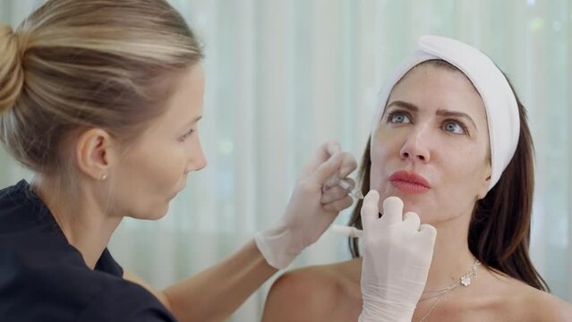 Surgeon, in medical gloves, carefully and slowly injects hyaluronic acid into woman's chin with a syringe. botox and filler augmentation procedure. beauty injections. Plastic surgery.