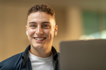 A young man with a beaming smile, using earbuds, and looking at a laptop screen