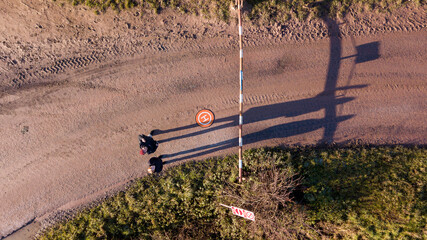 This aerial image captures a solitary figure at a crossroads, the long shadows of late afternoon stretching across the textured ground. The crossroads are marked by a traffic sign indicating a speed
