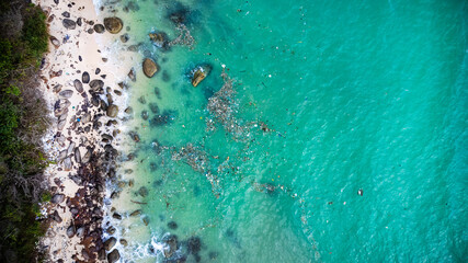 Aerial view of a tropical beach with crystal-clear water, sandy shore, and scattered rocks, depicting a serene vacation destination