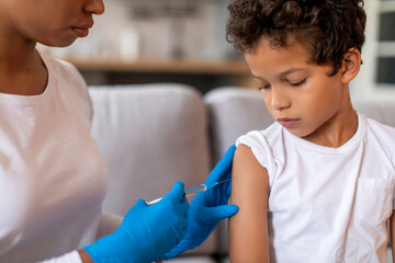 Doctor making vaccination against flu for little black boy in home interior - obrazy, fototapety, plakaty