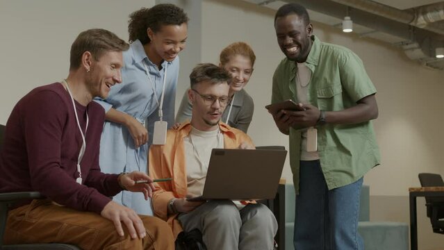 Team of multi-ethnic coworkers standing around businessman in wheelchair and discussing project on laptop with him during workday in office