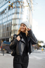Happy beautiful young woman model with a knitted hat and black leather jacket with a bag walking in the city