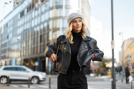 Stylish beautiful young woman in fashion urban clothes with a knitted hat, black sweatshirt, leather rock jacket walks in the city