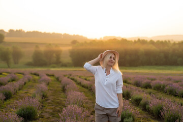 Beautiful young healthy woman with a white dress running joyfully through a lavender field holding a straw hat under the rays of the setting sun