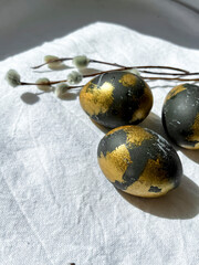 Dark blue and gold Easter eggs lie on a white light tablecloth under the rays of the sun, willow branches lie nearby. An Easter card. Vertical photo