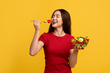 Healthy smiling asian lady eating fresh salad over yellow background