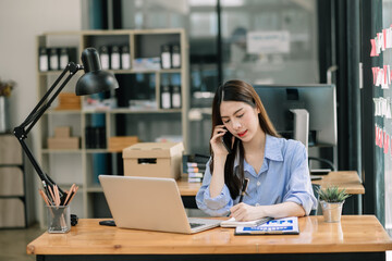 Young Asian businesswoman working with working notepad, tablet and laptop documents talking on the smartphone, tablet and laptop video call tax, report, accounting in the office