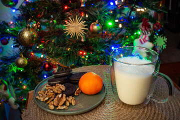 a plate with cookies, walnuts and a mandarin fruit next to a mug with milk with a defocused christmas tree in the background