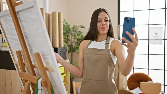 Charming young hispanic artist engaging in a joyful video call while crafting a beautiful drawing at her bustling art studio