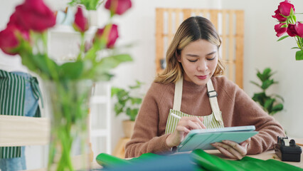 Small business concept. Young asian woman florist using tablet writing on order online at flower shop. Female florist or flower shop owner using tablet working, receiving order at flower shop