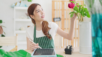 Small business concept. Young asian woman florist using tablet writing on order online at flower shop. Female florist or flower shop owner using tablet working, receiving order at flower shop - Powered by Adobe