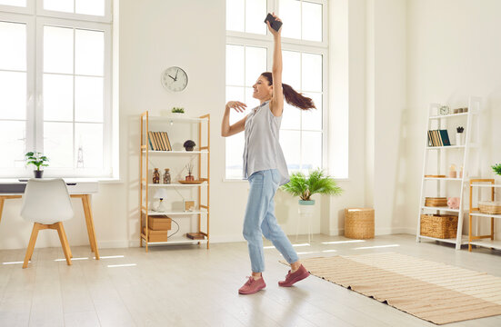 Full body photo of a young happy smiling girl wearing casual clothes dancing with smartphone in hand in the living room at home. Beautiful woman having fun with a satisfied face expression.