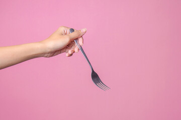 Woman's left hand holds a metal fork on a pink background.