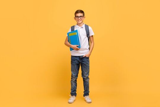 Portrait of smiling schoolboy with backpack and workbooks posing over yellow background