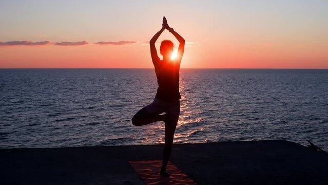Silhouette woman doing tree pose while exercising. Female is practicing yoga on seashore during sunset. She is meditating at beach.