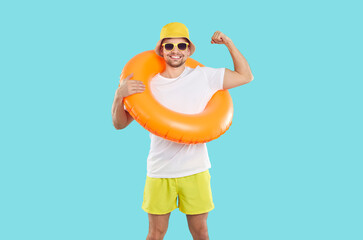 Cheerful man with inflatable swimming circle shows biceps on light blue background. Smiling male vacationer, humorous strong swimmer or beach lifeguard in T-shirt, shorts, panama and sunglasses.