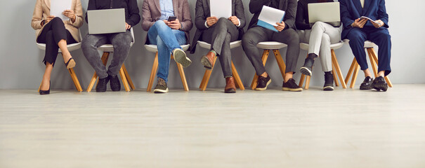 Group of people sitting in row in the office, waiting for a business meeting or job interview, working on laptop notebook PCs, holding clipboards. Cropped shot, human legs, banner background
