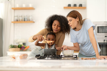Multiracial lesbian couple cooking healthy food in kitchen for their biracial boy. Gay women standing at counter enjoy preparing lunch at home with child kid. Young adult LGBT family eating together