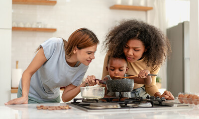 Multiracial lesbian couple cooking healthy food in kitchen for their biracial boy. Gay women standing at counter enjoy preparing lunch at home with child kid. Young adult LGBT family eating together - Powered by Adobe