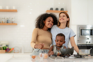 Obraz na płótnie Canvas Multiracial lesbian couple cooking healthy food in kitchen for their biracial boy. Gay women standing at counter enjoy preparing lunch at home with child kid. Young adult LGBT family eating together