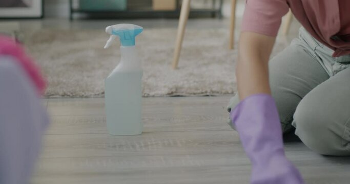 Close-up of female hand in rubber gloves spraying soap on wooden floor and scrubbing laminate doing housework. People and hygiene concept.