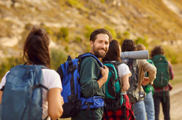 Team of friends trekking or hiking with backpacks. Happy man looks back at the photo camera while walking together with a group of people about a hilly rural area. Tourism, active lifestyle concept