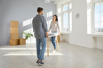 Happy family couple carrying furniture while moving into a new house or apartment. Joyful young man and woman carrying a white desk or table in an empty, unfurnished, light, modern living room at home