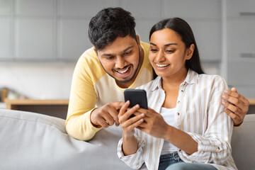 Cheerful Young Indian Couple Using Mobile Phone And Embracing Together At Home