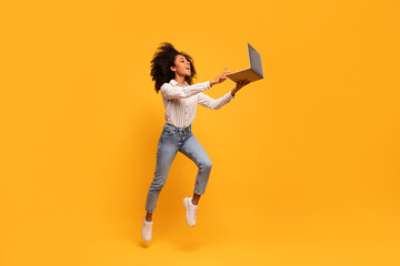 Energetic black woman leaping with laptop on yellow background