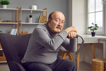 Portrait of a senior man sitting at home in rehabilitation with his crutch and looking sad at the camera in eyeglasses. Red haired elderly man with walking stick sitting on a cozy armchair.