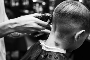 Black and white photo. Cute emotional fair-haired smiling boy with blue eyes at the barber shop. Stylist's hands with tools. Indoors, dark background, neon. Selective focus
