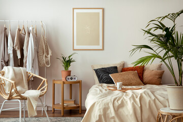 Creative composition of cozy bedroom with mock up poster frame, beige bedding, hanger with clothes, wooden bedside table, plants, pillows and personal accessories. Home decor. Template.