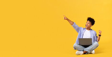 Joyful male student with laptop pointing at free space, sitting on yellow background