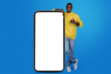 Joyful African American man in yellow hoodie giving a thumbs up next to a huge blank smartphone screen