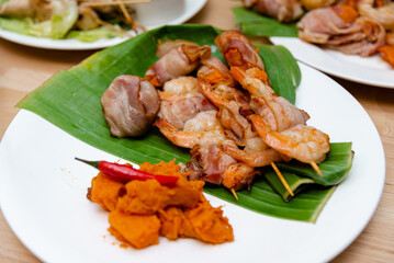 Bacon-Wrapped Shrimp and Grilled Meat on Banana Leaf. A delicious serving of bacon-wrapped shrimp alongside grilled meat and mashed sweet potatoes, presented on a banana leaf over a white plate.