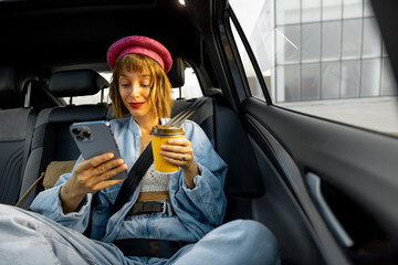 Young stylish woman uses smart phone while sitting relaxed with a coffee cup on backseat of car on...