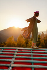 Woman enjoys beautiful sunset in the mountains, while standing on the roof of unfinished house....