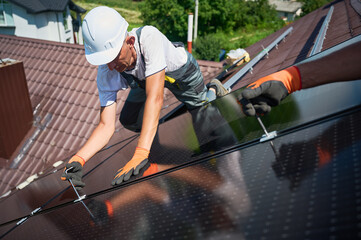 Workers building photovoltaic solar panel system on rooftop of house. Close up of men technicians in helmets and gloves installing solar module with help of hex key outdoors. Renewable energy.