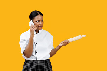 Concentrated black female chef holding rolling pin and talking on cellphone