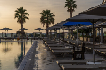 Sunrise over the pool and sunbeds. On the background of the sea coast. Big palm trees.