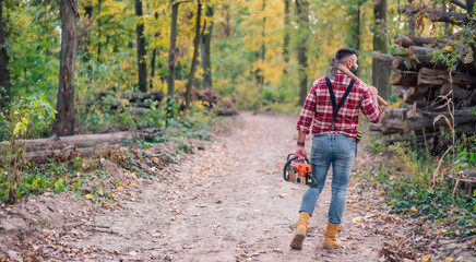 Portrait of a stylish hipster confidently navigating the forest path with an axe and chainsaw in...