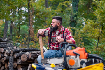 Caucasian man, a lumberjack, takes a moment to rest on a pile of firewood, pouring himself a cup of coffee from a thermos, his tools laid out in front of him.