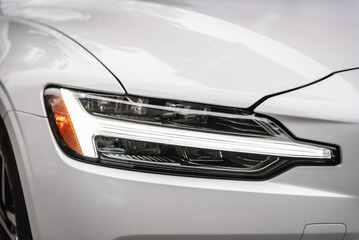 Front right headlight on modern white or silver new car. Exterior automobile closeup. Detail white metallic bumper car with light and car wheel. Car lamp signals to turn a car on the street.